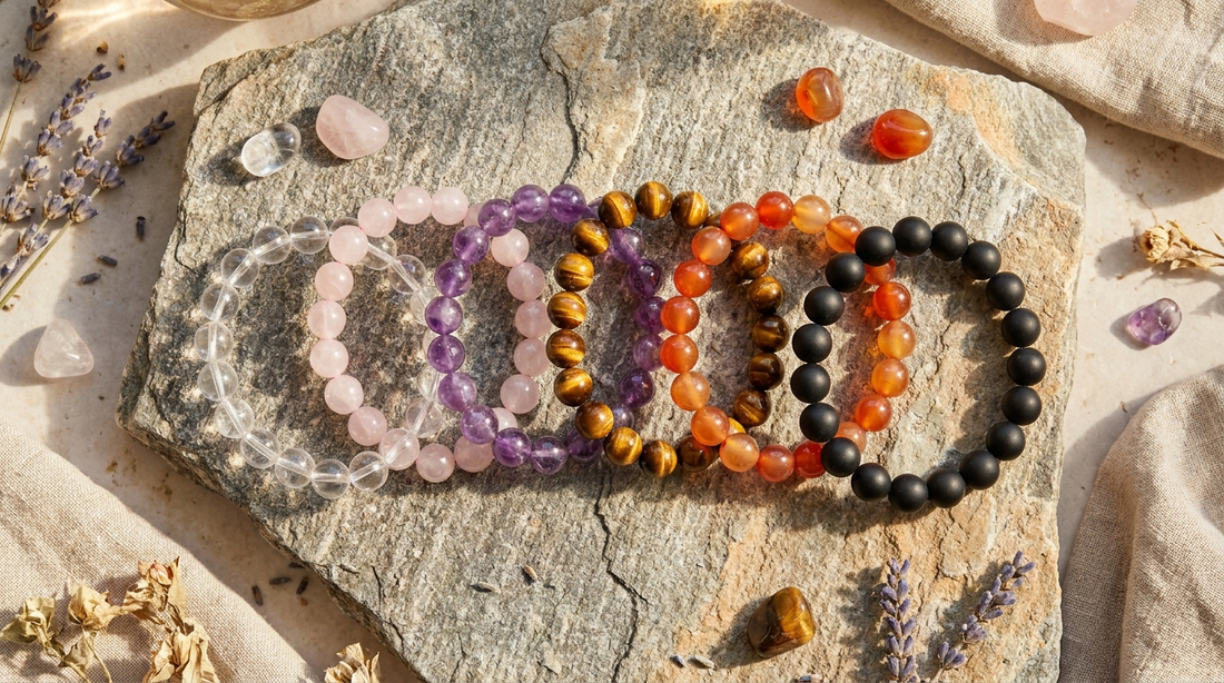 Six crystal bracelets — clear quartz, rose quartz, amethyst, tiger's eye, carnelian, and obsidian — arranged in a fan on a stone surface